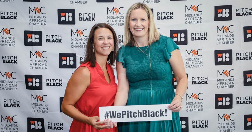 Two women - Katie and Leanne stand in front of the Pitch Black photo-op background