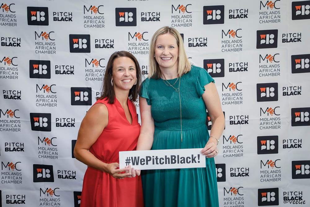 Katie and Leanne stand in front of a step and repeat holding a sign that says #WePitchBlack