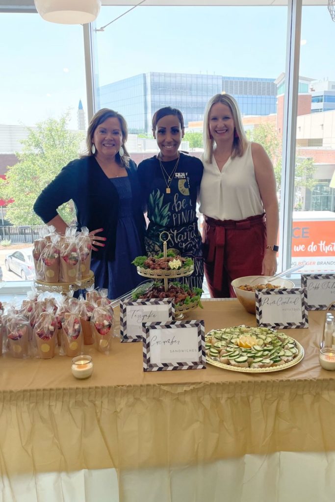 Three women pose with a catered lunch spread