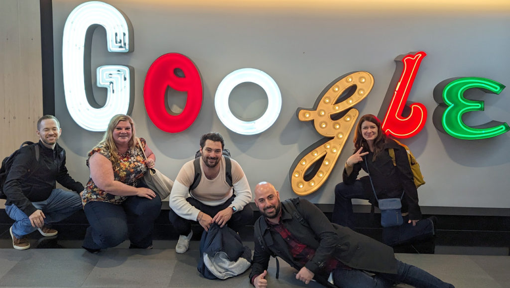 A group of five people posing in front of the Google sign at Google's offices in New York