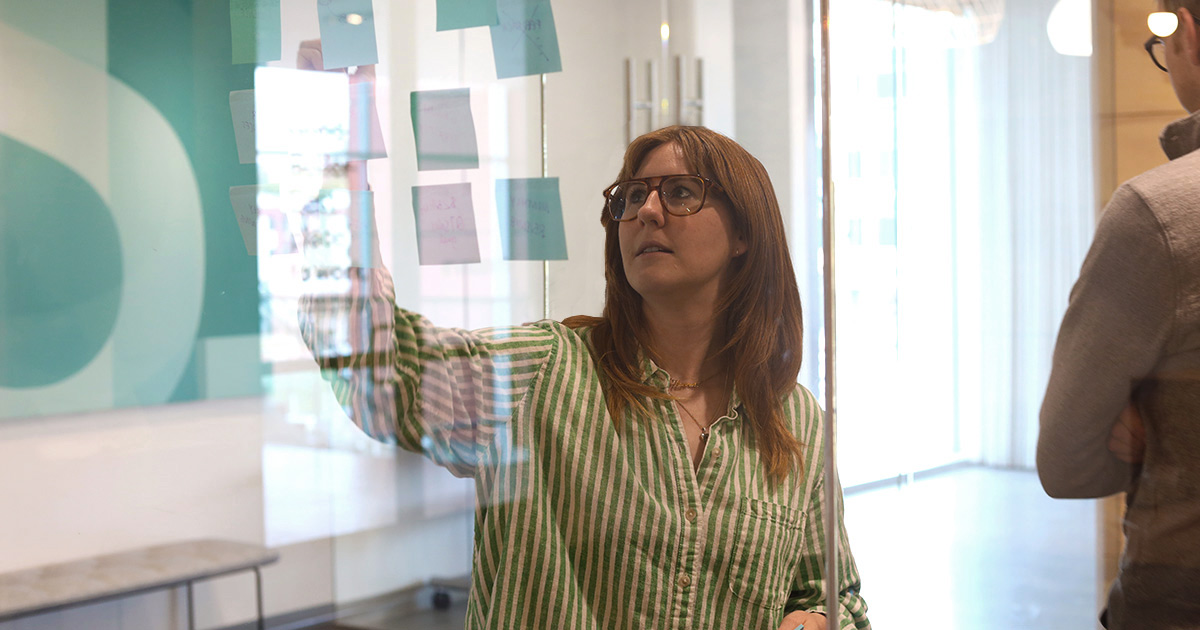 Woman looks at glass board with sticky notes that have different ideas written on them.