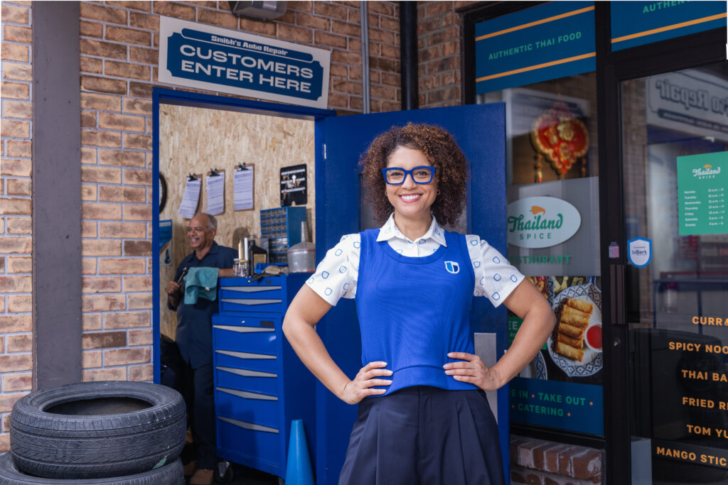 Berklyn, biBerk's spokesperson, smiling confidently with hand on her hips, standing in front of a strip mall with a mechanic and Thai restaurant.