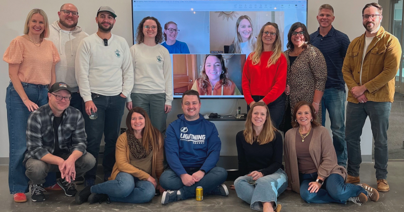 A picture of the E&S team in two rows. The front row is sitting and the back row standing around a monitor with three remote employees faces on it.