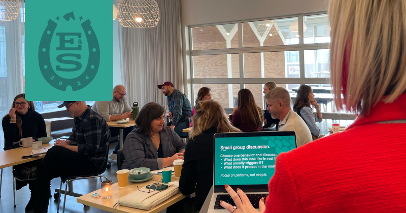 You see the back of a presenter wearing a red blazer. She is looking out at a room full of people sitting at tables of four.