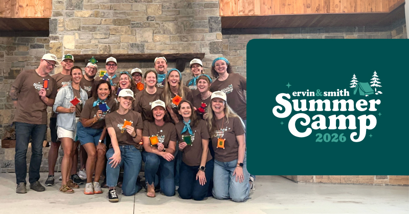Group of 20+ Ervin & Smith employees smiling at the camera. They are standing in front of a large stone fireplace.