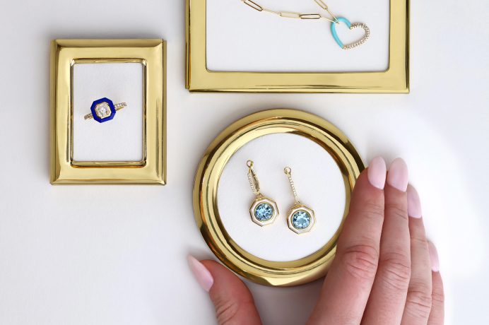 Image of three small gold metal frames. One frame has a pair of aquamarine earrings, one a diamond ring and the other a gold necklace with a heart pendant. There is a hand holding the frame with the earrings on it.
