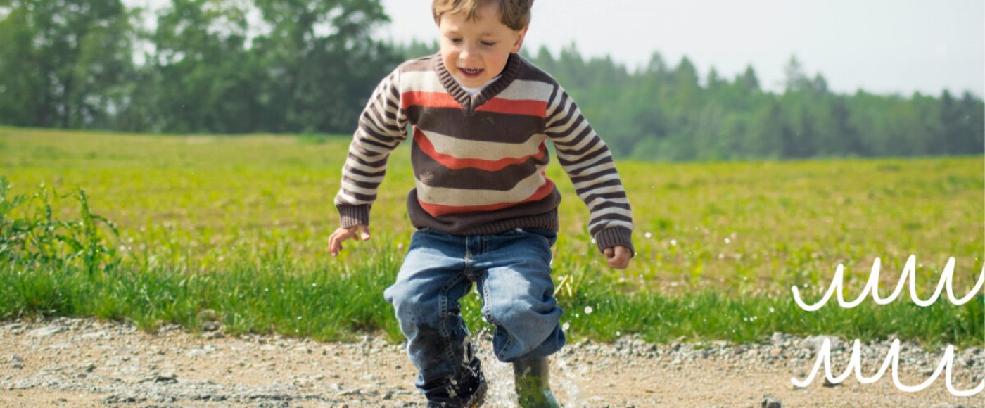 Happy, smiling boy is jumping in a puddle of water outside in a country or farm landscape.